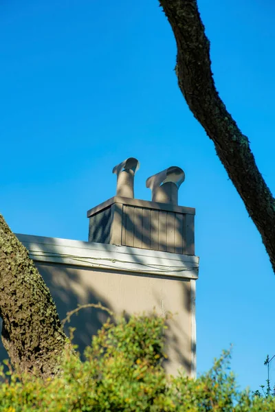 Metal chimney with beige stucco exterior and pipes on top with front yard trees and shrubs with shadow and some sun light. In the suburban area of the downtown neighborhood for house details on building.