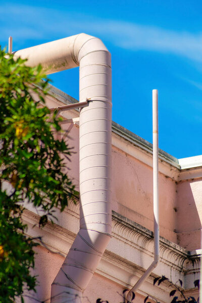 Pink ventilation pipe in partial sun and shade with foliage or trees in the yard on building with blue sky background with clouds. In the city or in a suburban area in the neighborhood.
