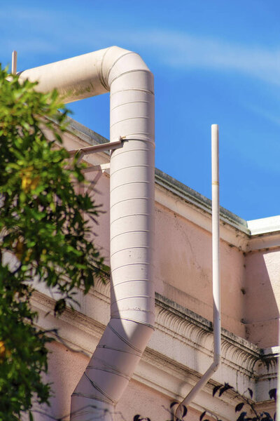 Pink ventilation pipe in partial sun and shade with foliage or trees in the yard on building with blue sky background with clouds. In the city or in a suburban area in the neighborhood.