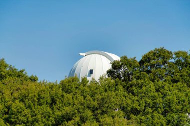 Güneş altında tepeleri ve teleskopu olan uzak beyaz gözlemevi kubbesi. Ağaçlı ve yapraklı. Ya da öğleden sonra mavi gökyüzü arkaplanlı.