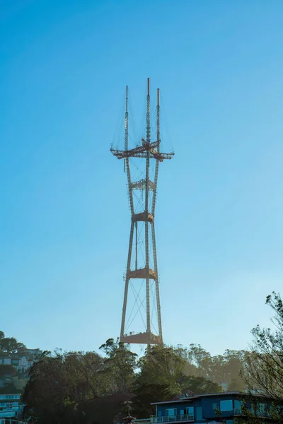 Sutro tower in the red and orange sunlight in the downtown districts of san francisco california in late afternoon shade in city. Blue and white gradient sky background with front yard trees in town.