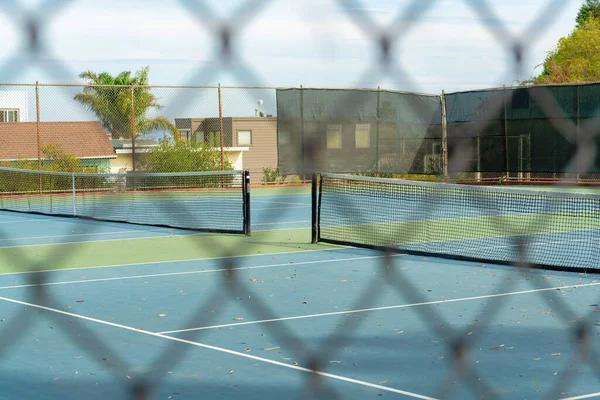 Blue and green tennis court with nets as seen through blurry metal chain link fence foreground in late afternoon shade. Sports court in the park in the city or neighborhood on cloudy day with trees.