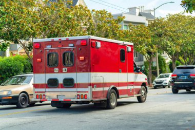 Şehir merkezindeki caddelerde hızla ilerleyen kırmızı ambulans arka planda ağaçlar ve yapraklar ve ön planda diğer arabalarla birlikte. Öğleden sonra güneşli bir günde acil bir duruma yardım etmek için..