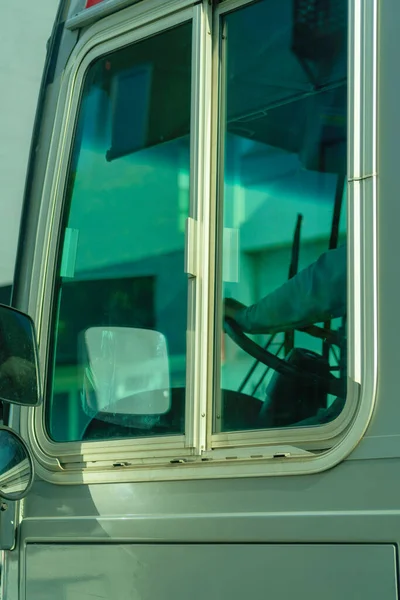 Bus window with blue tint on the glass and gray metal panneling on the side of car. In late evening sun for public transportation in the city and a visible steering wheel.
