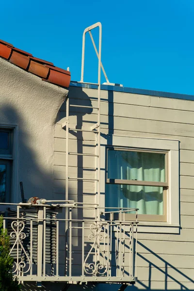 Beige metal fire escape ladder on side of house or bulding with visible window and blue sky background. Late in the afternoon with red roof tiles and balcony for saftey and escape.