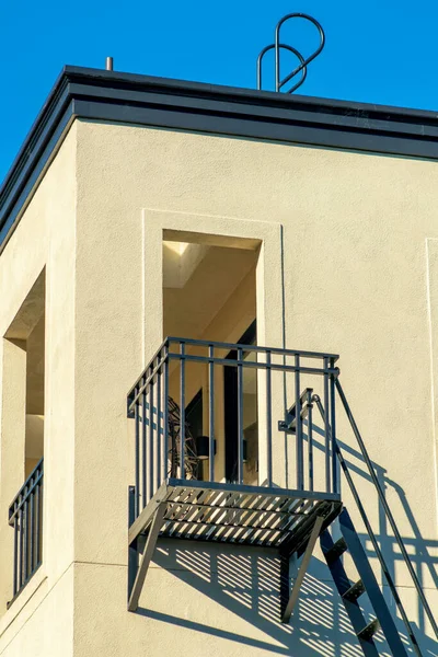 Lone balcony with fire escape ladder and rooftop access with beige stucco exterior with blue sky clear background. Late afternoon sun with visible opening windows and dark flat top.