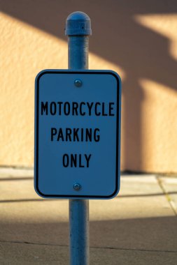 Black and white road sign that say motorcycle parking only in late afternoon shade with urban backdrop stucco wall. In the city for transportation and storage or bikes or cars in a dense urban area.