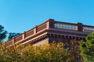 Flat roof balcony with decorative hand rail facade on exterior of orange or red building or structure in midday sun. Visible front yard trees with clear blue sky in the downtown city neighborhoods.