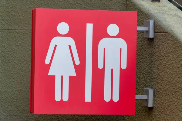 Red bathroom sign with woman and man pattern in white paint on metal post indoors with beige stucco wall in shade. In the city in a building for using restroom with toilet sink and urinal.