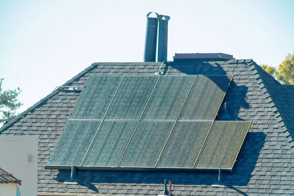 Dark solar pannels on top of steep roof on house or home with gray roof tiles and metal chimney vents in suburbs. Clear blue sky on residence in downtown city neighborhood in the city.