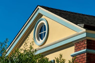 Nautical round window in attic room at pinnacle of home with beige stucco exterior and gable style roof with white accent. Brick with green trim and fornt yard trees with clear blue sky background.