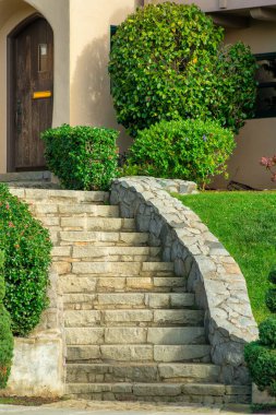 Sprawling stone staircase bending down steep front or back yard near home or residence entrance on house in neighborhood. Green lawn and shrubs near brown wooden front door.