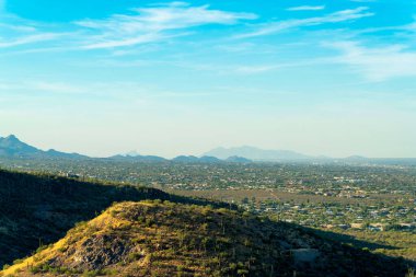Hillside mounds in the moutains of Tuscon Arizona in late afternoon with suburban and urban neighborhoods. Blue sky with clouds and cactuses and native plants and foliage in distance.