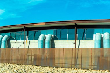 Exterior of water processing plant with blue pipes leading from building and cement walls with metal tin or titanium roof. In late afternoon sun with front or back fence in an industrial part of city.