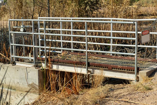 Metal footbridge in industrial area to cross over to hand wheele valve to turn on and off the flow of water in sun. Water dam or facility for conservation purposes with visible grass in sun.