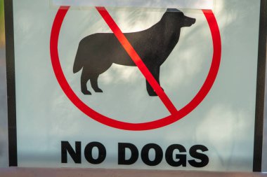 Sign that say No Dogs with a black dog silhouette and a red line through the dog symbol on white paint with black lines. In shade to show that k9s are restricted from a given park or recreation area.