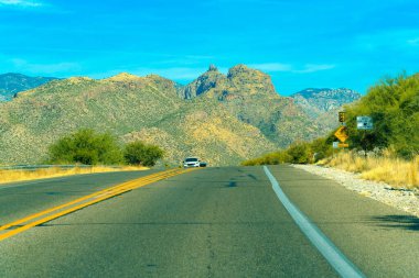 Road curving over a ridge in the hills of Arizona in the heat of the midday sun with visble cars and lines and blue sky. Moutains in distance with natural cactuses and foliage in north america.