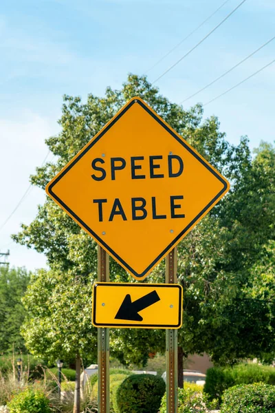 Road sign that says speed table with black and yellow paint on exterior in late afternoon shade in summer sunlight. Transportation symbol to tell drivers to slow down in a neighborhood.