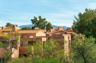 Row of modern apartment buildings with adobe style roofs and exteriors with beige stucco and orange tiles. In late afternoon sun with hazy blue sky and front yard trees in desert neighborhood.