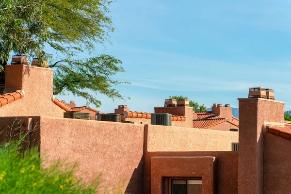 Adobe style stucco exterior with air condition units on flat roof with visible chimneys and desert style architecture. In late afternoon sun with back and front yard trees in neighborhood.