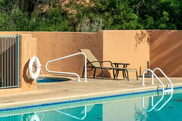 Pool side view with orange stucco wall and metal hand rails with water and cement with saftey float in midday sun. Pool in apartment complex or community public area used for swimming and recreation.