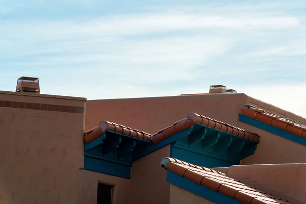Multiple roof facades with ledges and visible chimney with blue and hazy white cloudy sky in late afternoon shade. Building on house or home in a downtown adobe neighborhood in city.