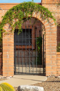 Brick archway with green vines on key stone with black metal gate in courtyard in suburban house or home front. In late afternoon sun with sidewalk and plants with gravel in rock garden.