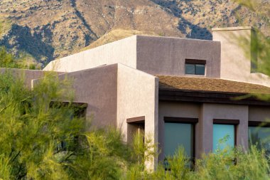 Adobe style house with flat roof and block features with visible windows and chimneys in the late afternoon sun and shade. Front yard trees and shrubs with white stucco exterior nearby moutians.