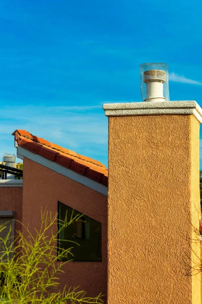 Chimney on modern house with slanted roof and red adobe tiles in late afternoon sun with blue sky and white clouds. In a suburban neighborhood with front yard trees in shade in desert community.