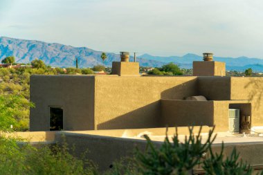 Adobe style building as house or home in the desert landscape with visible cactuses and trees in wilderness. Chimneys and windows on structure with mountain background and blue and white hazy sky
