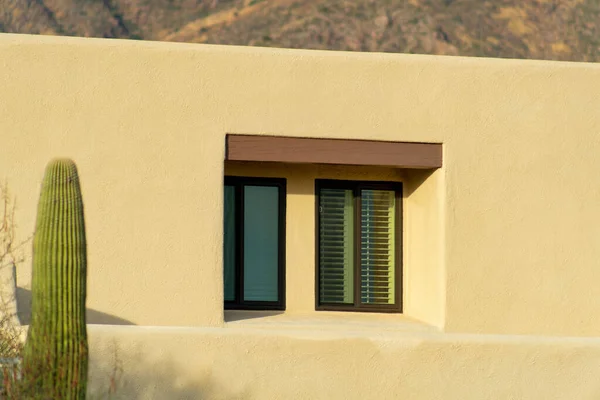 Adobe style building with view of small window and moutains in background with small saguaro cactus in front yard. Stucco cement exterior of building in late afternoon sun in the desert ladscape.