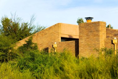 Brick building exterior with chimneys and visible windows on side of house or home in desert landscape in sunset with cactuses. In rural area with natural grasses and trees with hazy white sky in sun
