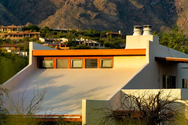 Modern house facade with white stucco and wood exterior and orange accent paint with slanted flat roof and windows. In late afternoon or early morning sunlight with mountain and natural background.