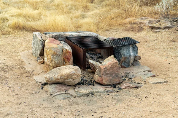 Black metal fire grate or pit with stabilizing rocks on sides in camp ground or near national park for barbeque and cooking. Late in the day with natural grass and dirt background for outdoors.