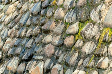 Close view of rocks building into a dam retaining wall used to control water levels and direct liquid from mountain run off. In late afternoon shade with protruding rocks and river moss on exterior.