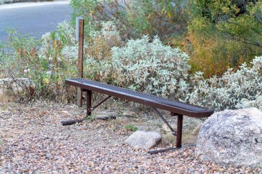 Metal bench with plastic seat and visible rocks and boulders with plants and shrubs with vegetation in background and shade. In late afternoon near side of walking road or path in great outdoors.