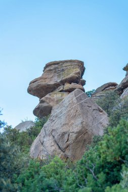 Rock formation with green trees at base in the cliffs and hills of the arizona desert in mountain region during dry season. Late afternoon shade with hazy white and blue sky with great outdoor stones.