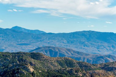 Rolling desert hills with powerful towering ridges and mountains in the cliffs of arizona desert in late afternoon sunlight. Day with hazy blue sky and clouds in great outdoors with trees and nature.