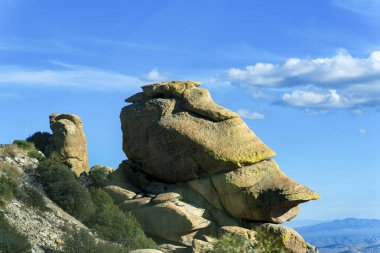 Tall rock formation on the edge of cliff or overhang on the top of mountain in afternoon sun with blue cloudy sky background. In the hills of arizona midday with vegetation and trees nearby.