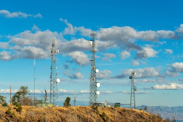 Row of industrial radio towers on summit of mountain in arizona with cloudy desert skies and blue with dry grass. Hills in industrial area for internet connection and radio and cell phone connection.