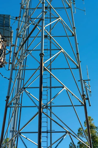 Radio or internet tower in industrial area with satelite dishes and triangular metal facets in afternoon shade with blue sky. Small access ladder for climbing to top and no clouds in afternoon sun.