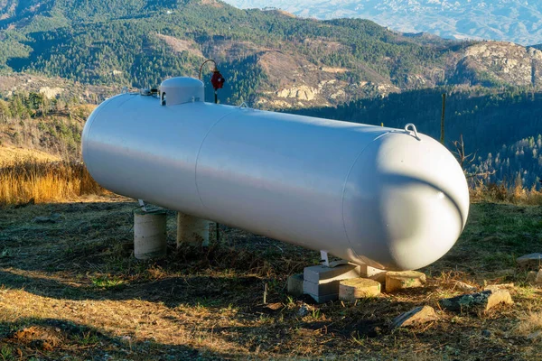 Metal round propane or natural gas tank in afternoon sun and shade on top of mountain or summit with rolling hills. Late in the day with white color paint and metal legs and tubes on body.