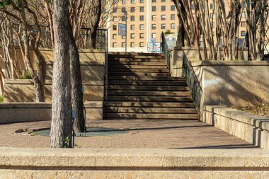 Path or walkway area with trees and cement wall with urban building background and sunny shade for resting weary travelers. Near park or sidewalk with bench and garden area to feed birds in town.