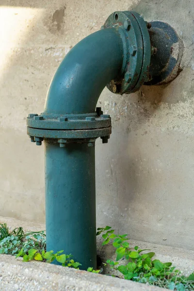 Blue green industrial sewage pipe on endge of building or structure with rivets and screws and small grass near sidewalk. In city or in urban area in late afternoon shade for plumbing and water.