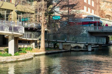 Canal in san antonio texas with pedestrian walkway and sidewalk and bridge for bus and traffic over river or waterway. Late afternoon sun and shade in business district with restaurants and shopping.