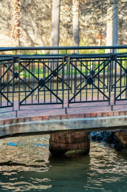 Köprü üzerinde tuğla kaldırım ve gölde yuvarlak beton direkli siyah metal parmaklıklar ya da gölgeli nehir kanalları. Doğanın kentsel ya da banliyö parklarında arka planda güneşli ağaçlar.