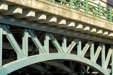 Massive archway bridge with metal beams and poles with cement and wooden fastening material for transport of vehicles. People walking on bridge with green hand rails to traverse river.