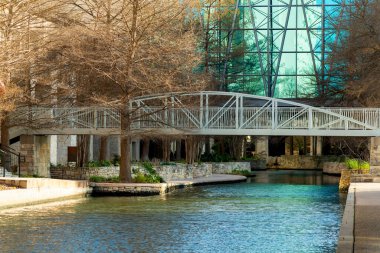 Metal bridge over canal or lake in urban or commercial district in mall or outdoor shopping center in san antonio texas downtown. Reflective glass building in afternoon sun with sidewalks.