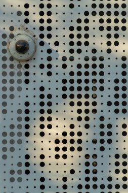 White hole punched metal building facade and exterior with decorative pattern and design with visible security camera. In late afternoon or early morning shade with round lense on side.
