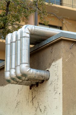 Vents bending out of building onto roof of stucco white and brown facade in urban or industrial area of neighborhood city. Late afternoon shade with trees in downtown suburban town.